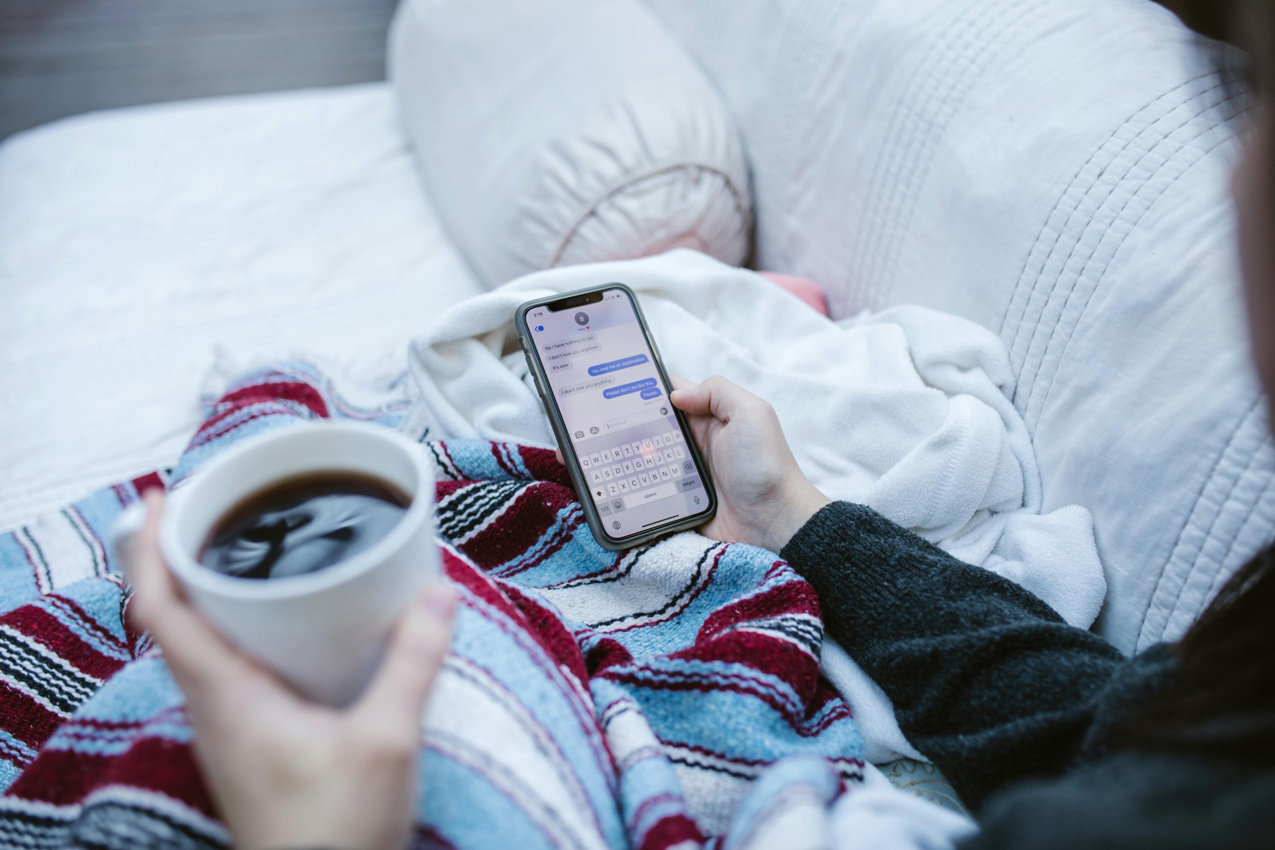 Person relaxing indoors with coffee and smartphone, texting in a cozy setting.